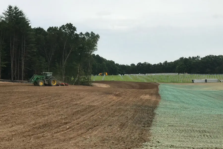 A field divided into tilled earth and green hydroseed, with a tractor, rows of stakes, and woods in the background.
