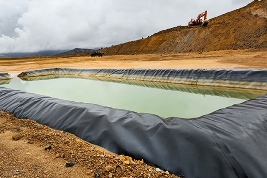 A geosynthetic-lined pond with greenish water in an industrial or construction area, with an excavator on a dirt hill.