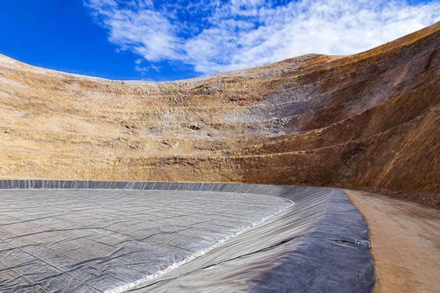 A massive open-pit mine or excavation with terraced walls, featuring a large gray HDPE liner at the base under a blue, cloudy sky.