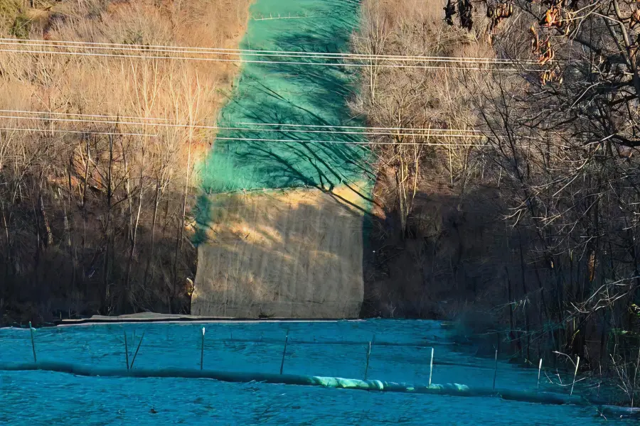 A steep hillside treated with green and tan hydraulic erosion control materials, flanked by bare trees and crossed by power lines.