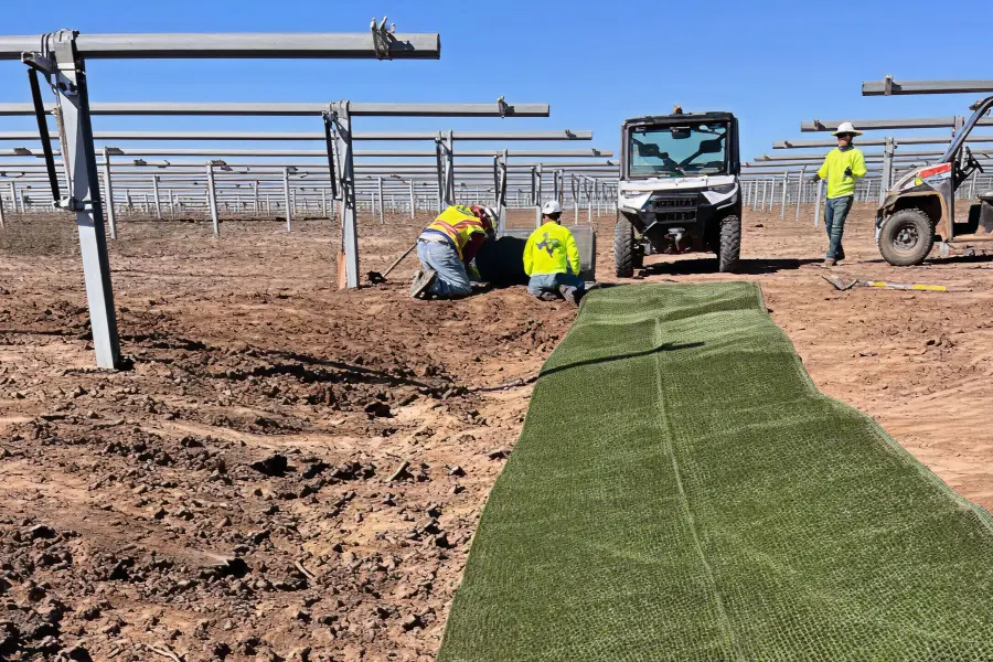 Workers install green erosion control mats at a solar farm construction site, with metal solar panel frames extending into the background.