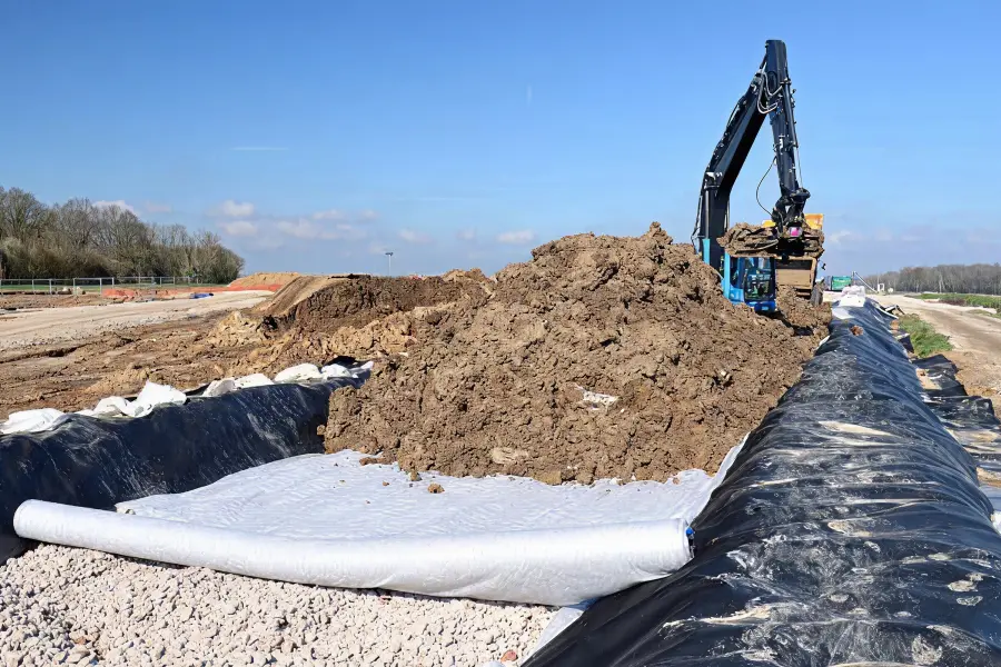 Excavator moves dirt on a construction site with black lining and white geotextile fabric being installed.