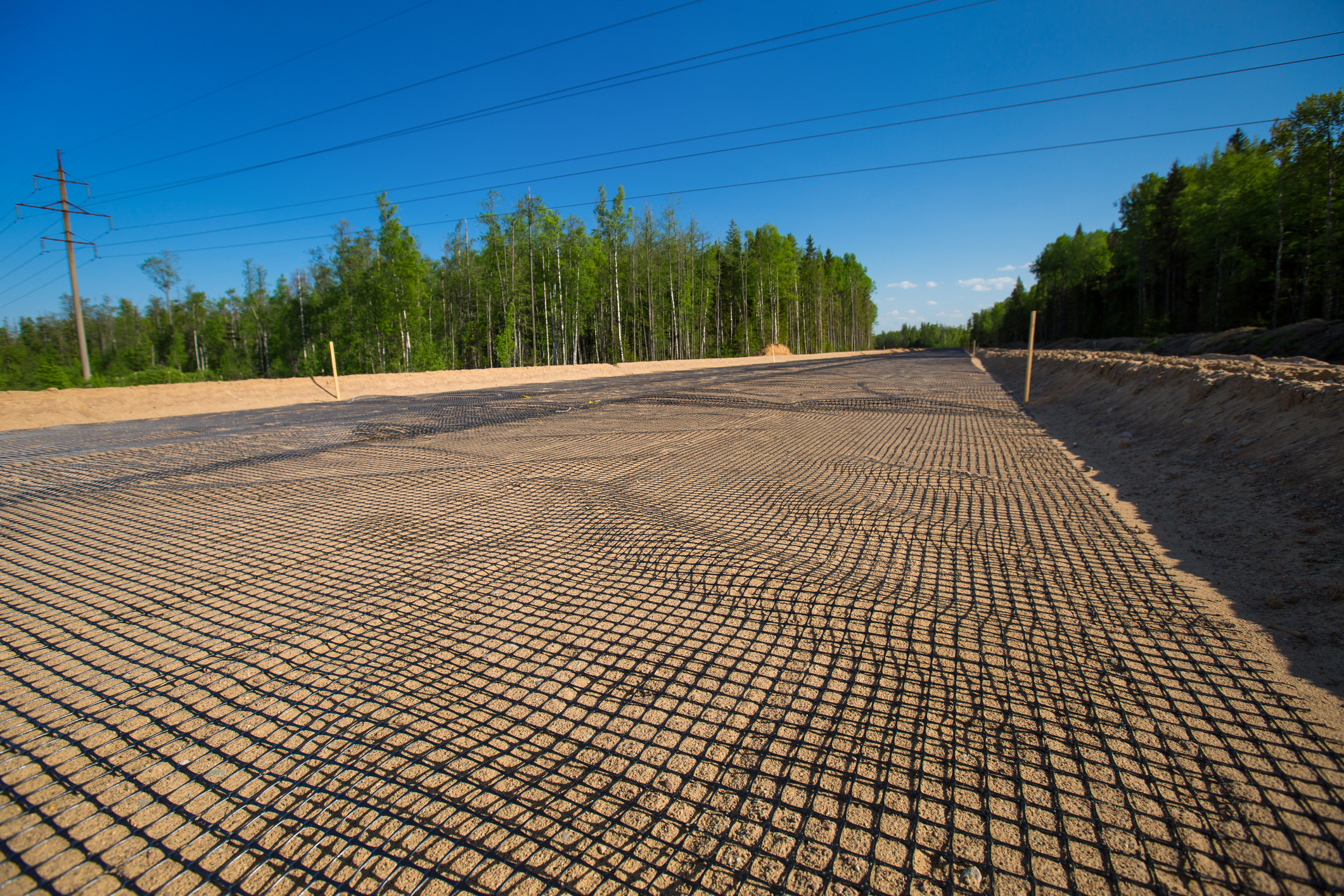 Geogrid reinforcement on a dirt road path at a construction site, with a distant forest and power lines under a clear blue sky.