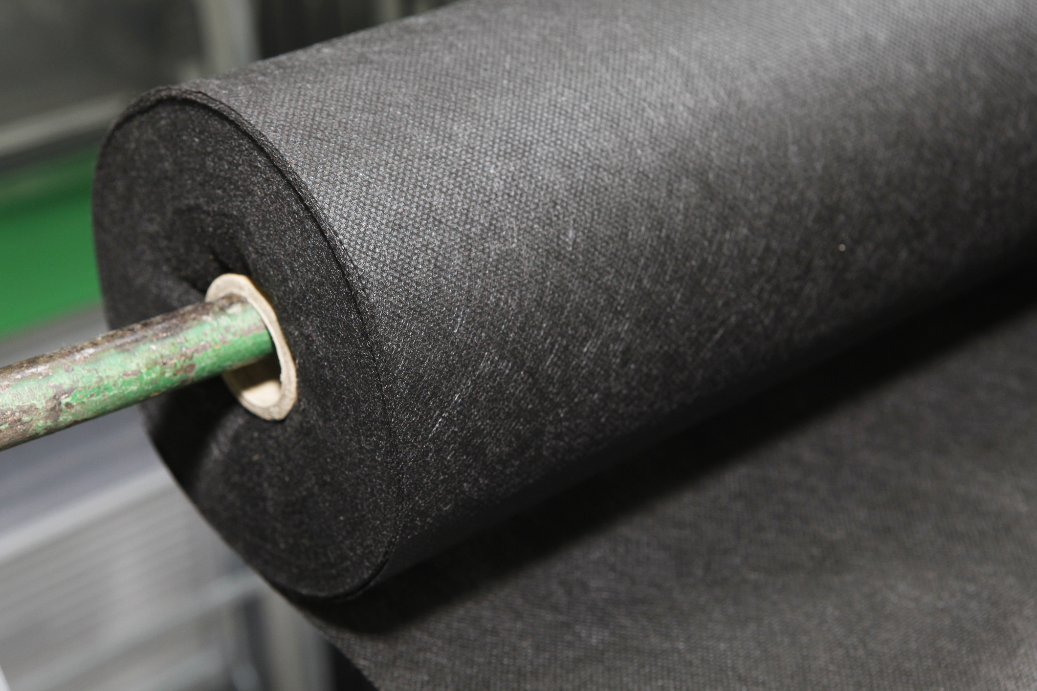 Close-up of a large roll of black non-woven fabric, likely geotextile, mounted on a green metal bar.