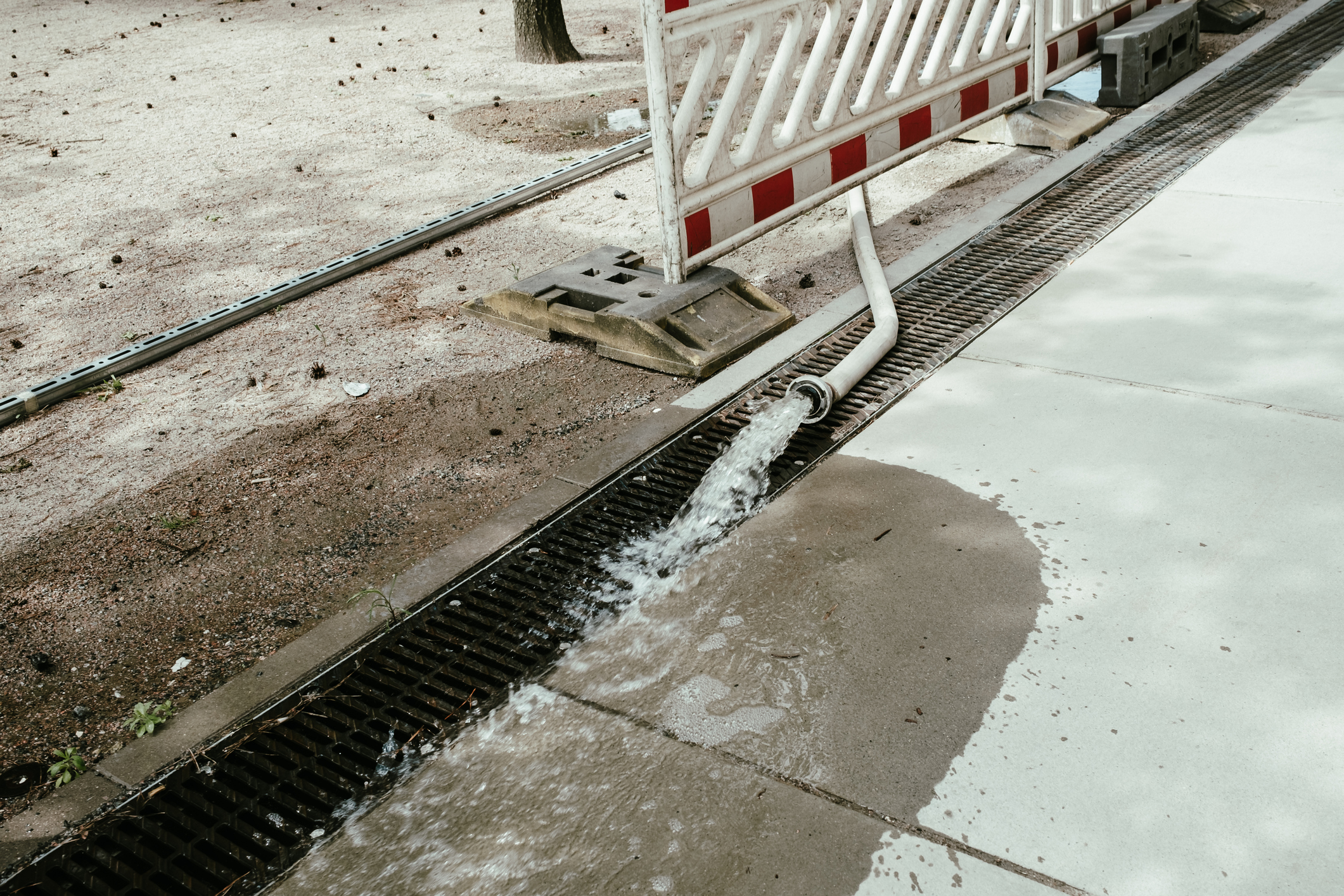 Water flowing from a hose into a grated strip drain on pavement next to a construction barrier and sandy ground.
