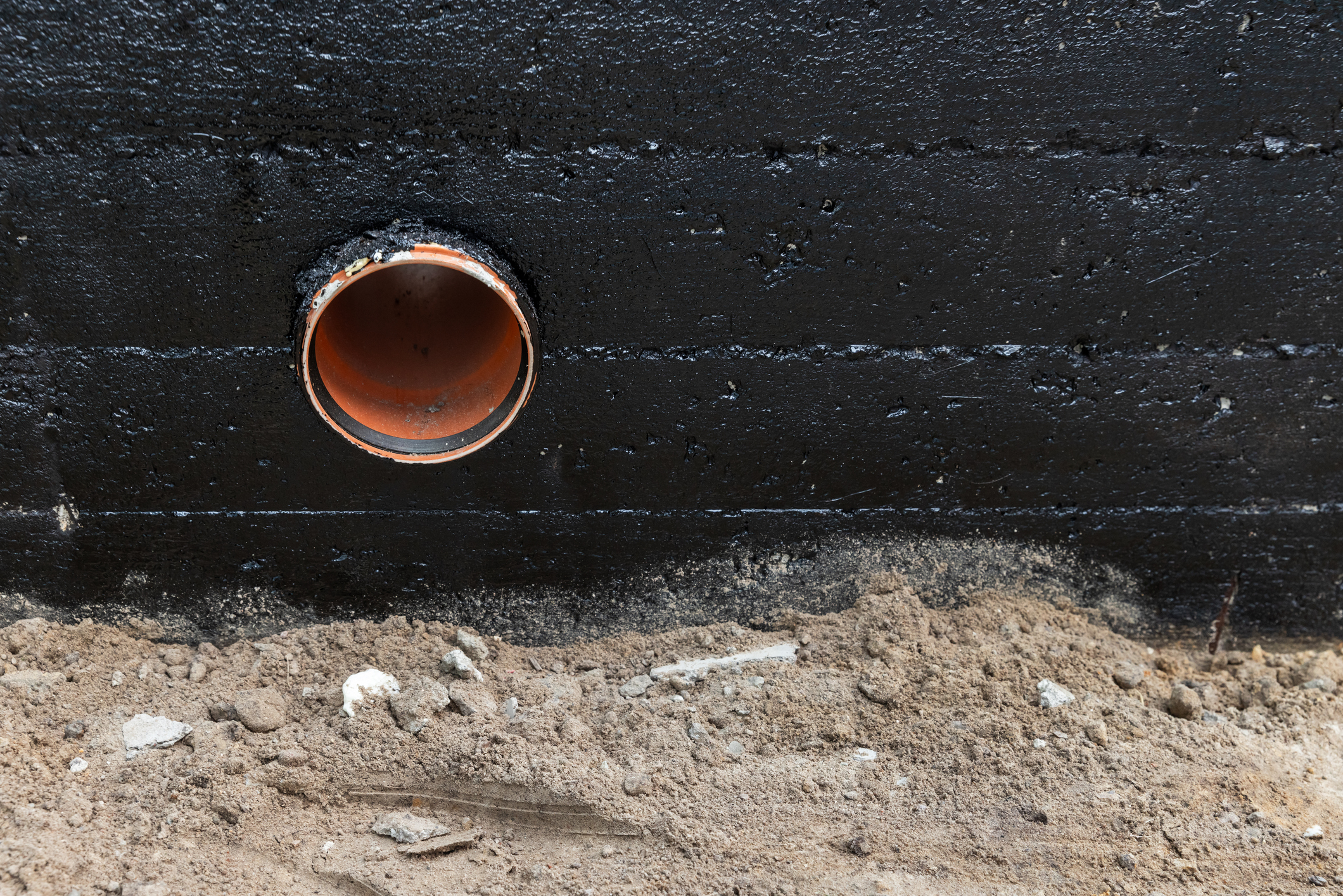 Orange pipe exiting a black waterproofed foundation wall, with sandy soil and construction debris below.